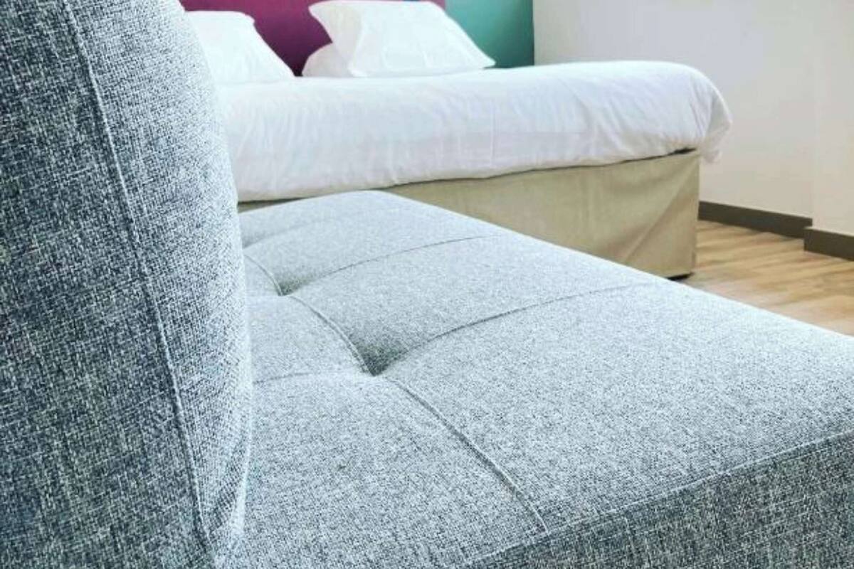 Een moderne slaapkamer met een grijze bank, een bed met wit beddengoed, en een raam dat voor natuurlijk licht zorgt.