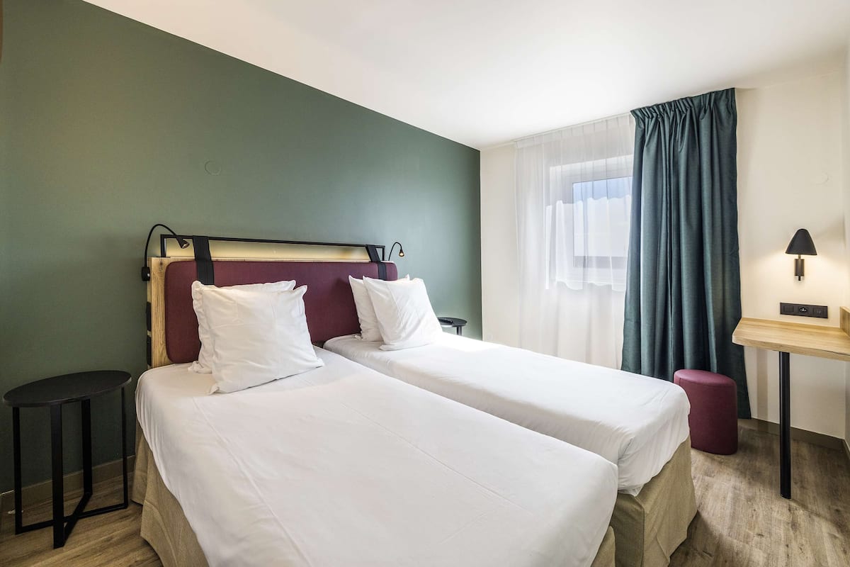Een hotelkamer met twee bedden, een klein tafeltje, een lamp en een raam met gordijnen.