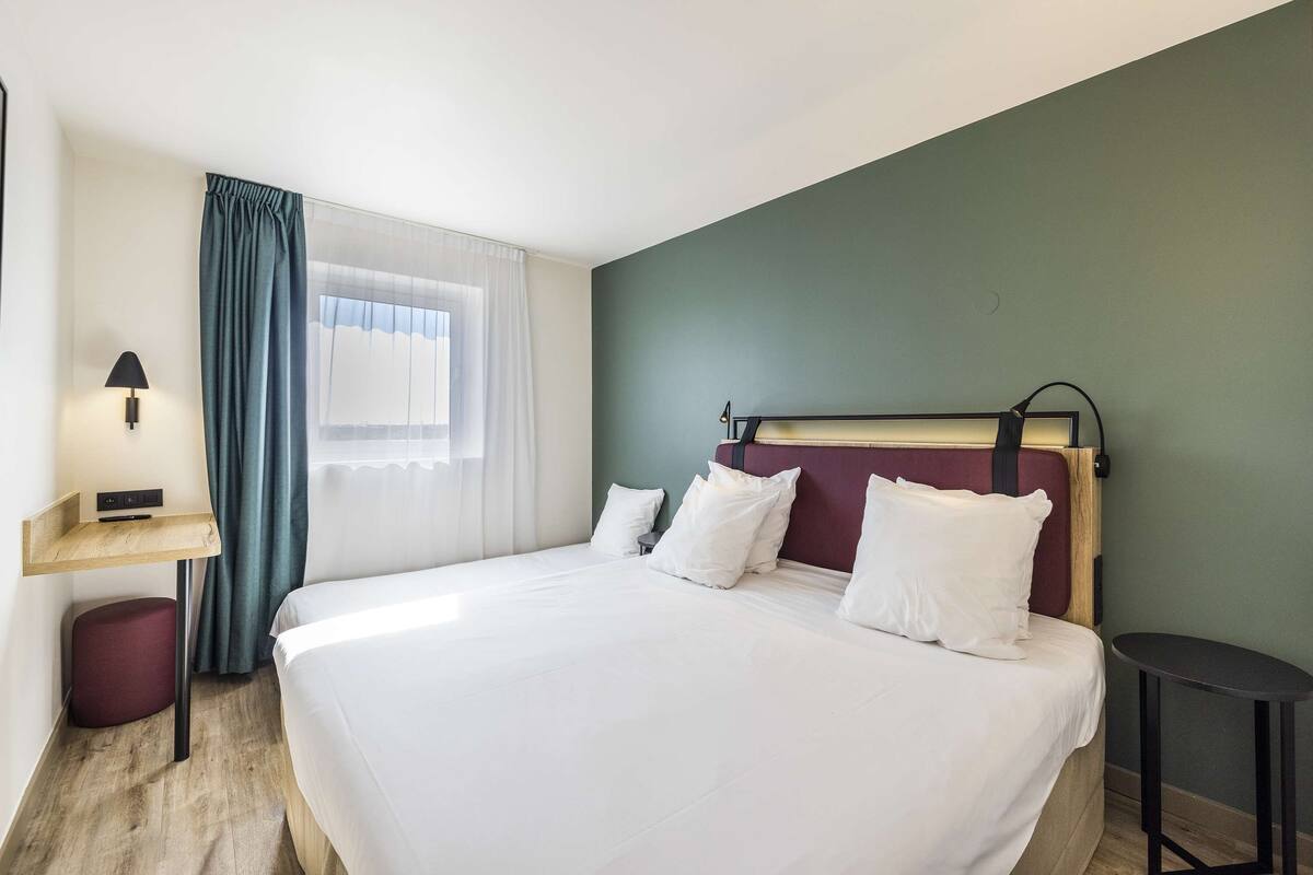 Een hotelkamer met een groot bed, een nachtkastje, een lamp, een raam met gordijnen en een klein krukje.