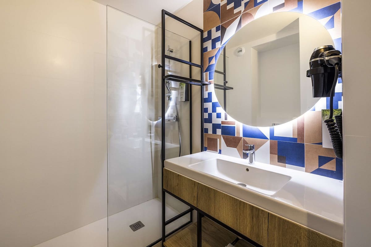 Een moderne badkamer met een douchecabine van glas, een wit wastafelmeubel en een spiegel met een geometrisch betegelde achterwand.