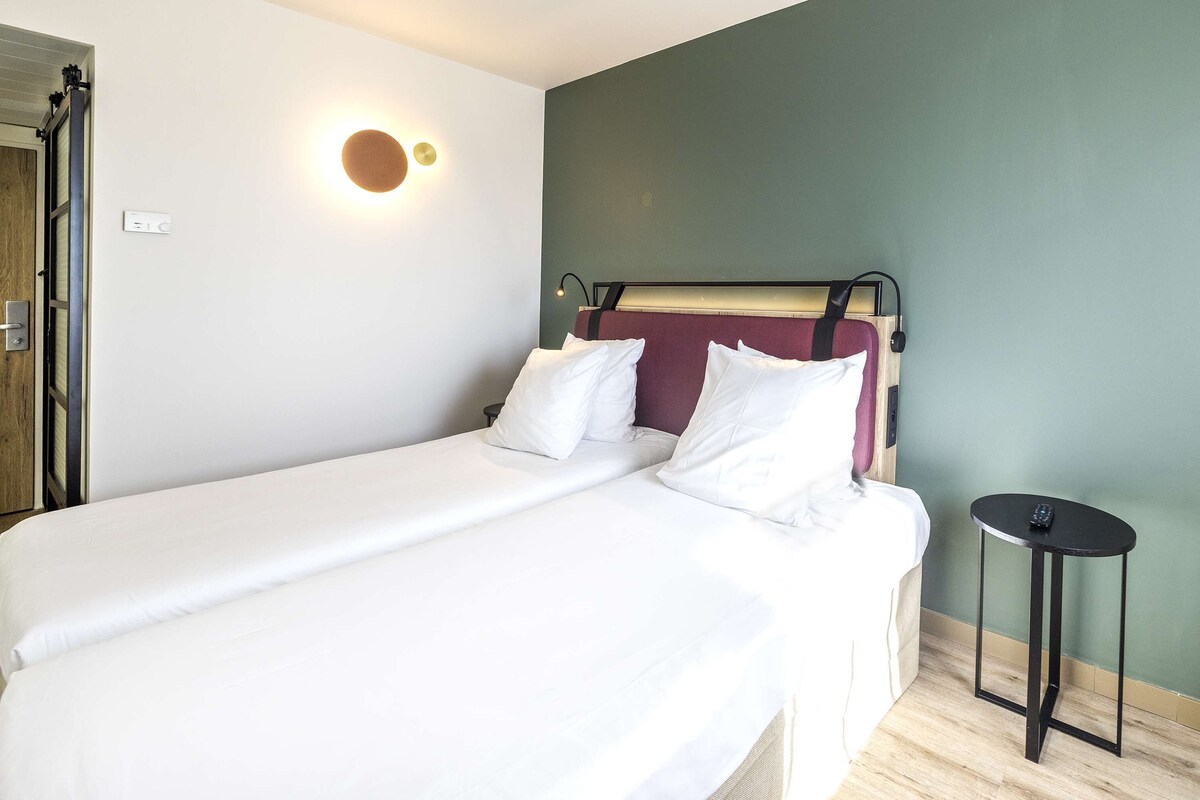 Una cama bien tendida con sábanas blancas, una lámpara de pared redonda y una mesita de noche negra al lado.