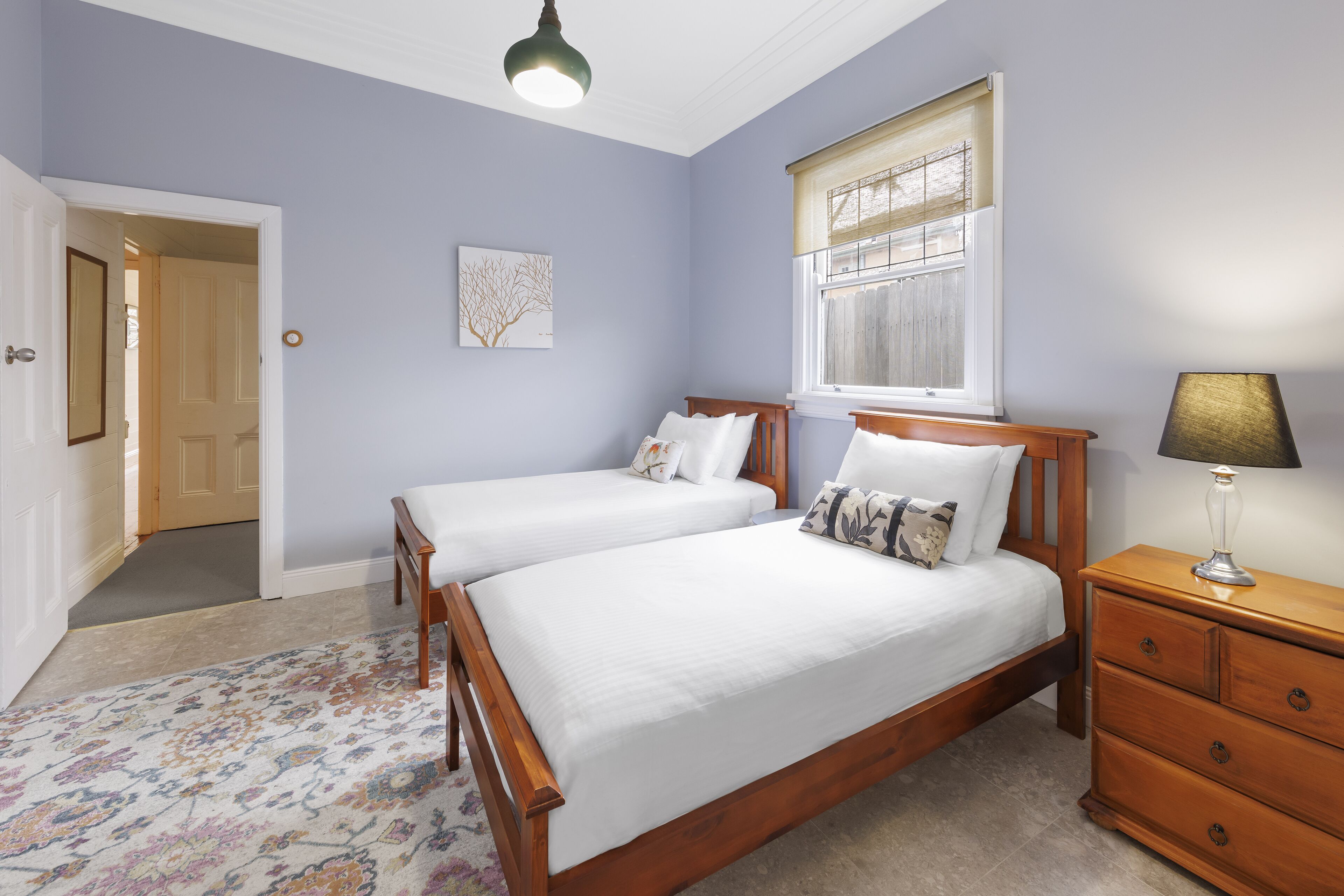 A bedroom with two wooden beds, a nightstand, a lamp, a window with blinds, a door, and a carpeted floor.