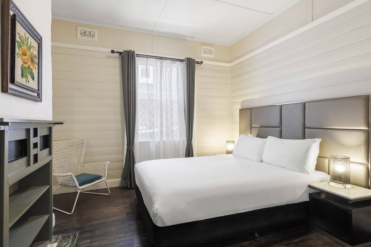 A neatly arranged hotel room with a large bed, a chair, a bedside table with a lamp, and a wall-mounted artwork.