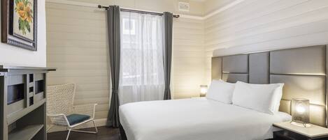 Camera d'albergo ordinata con un letto grande, una sedia, un comodino con una lampada e un'opera d'arte appesa al muro.