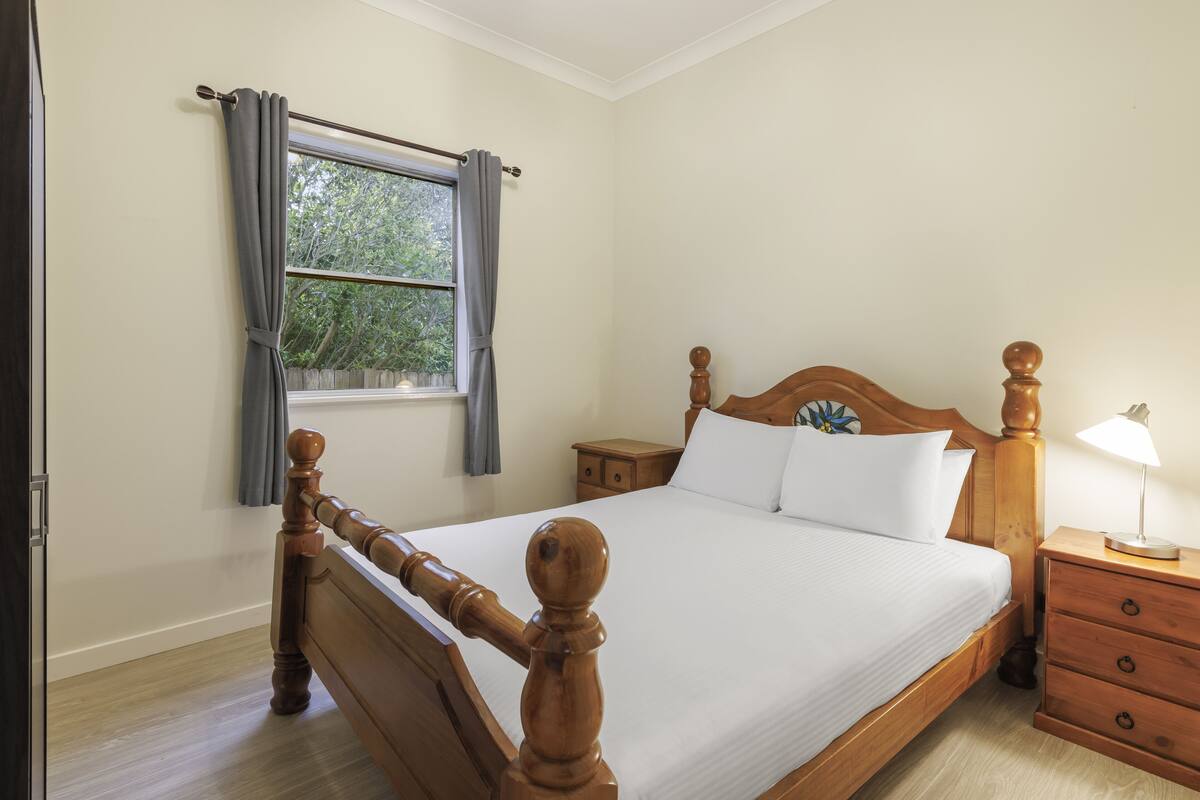 A neatly made bed with a wooden headboard, a bedside table with a lamp, and a window with curtains.