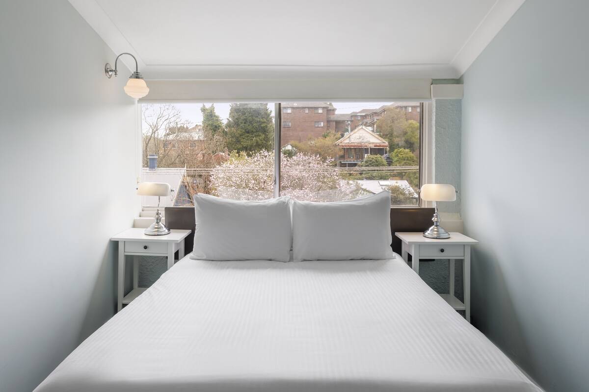 A bedroom with a large bed, two bedside tables with lamps, and a view of a garden and buildings through a window.