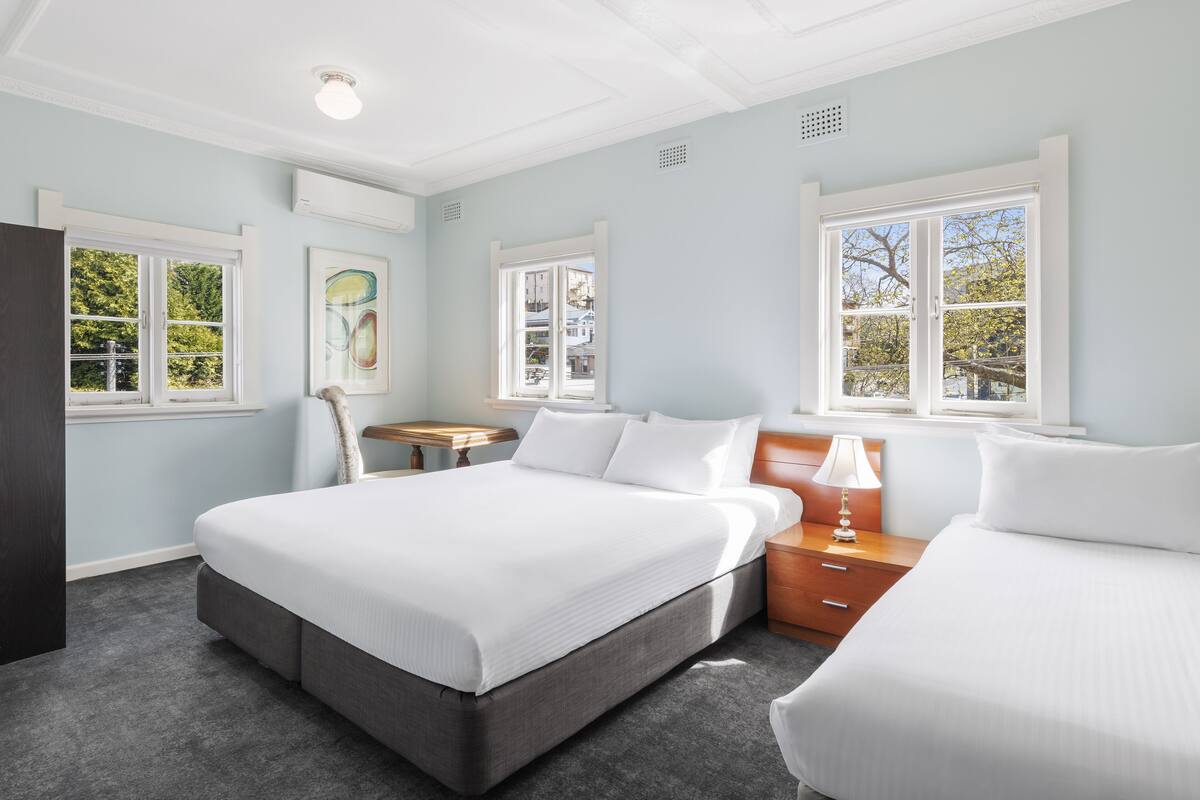 A hotel room with two beds, a wooden nightstand, a lamp, and a view of trees through the windows.