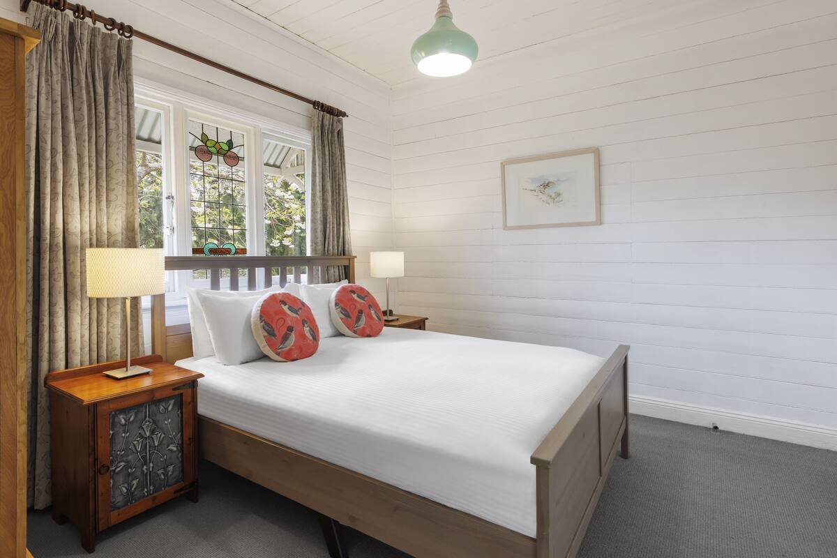 A neatly arranged bedroom with a wooden bed, a nightstand with a lamp, a window with curtains, and a wall-mounted picture.