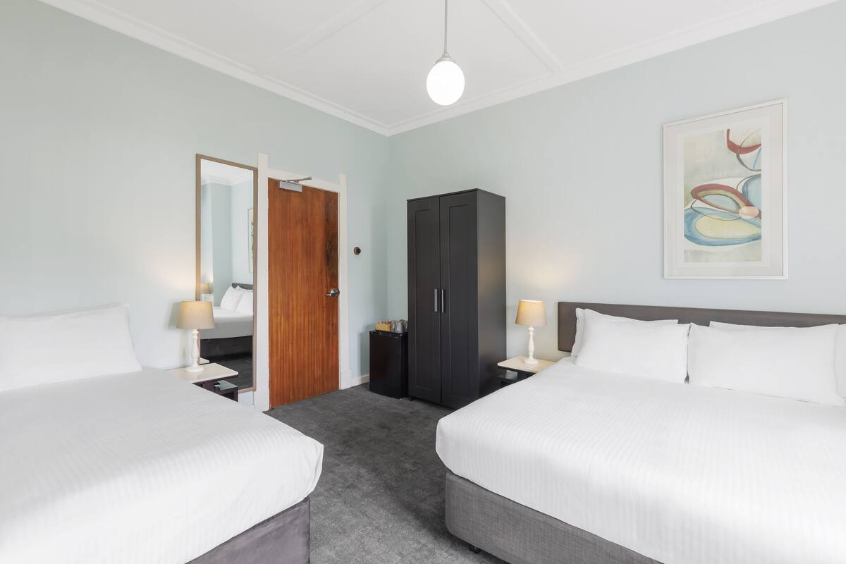 A hotel room with two beds, a wardrobe, a mirror, and a framed abstract painting on the wall.