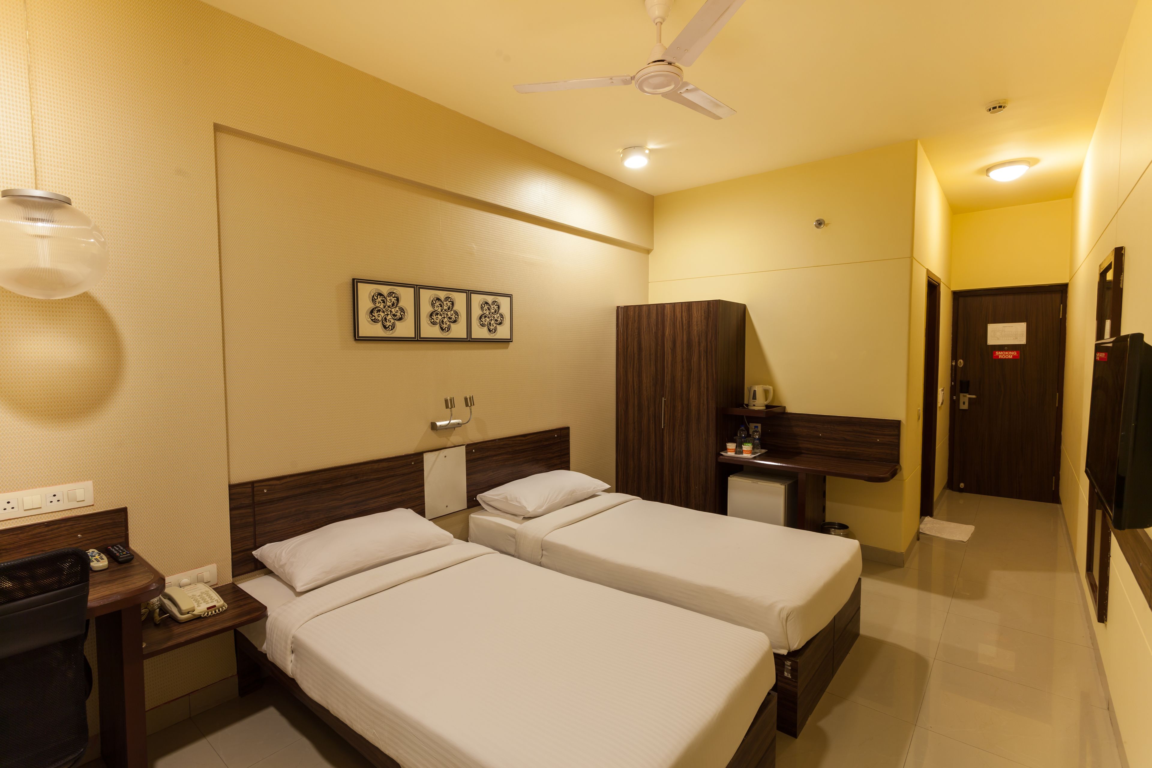 A hotel room with two beds, a desk, a wardrobe, and a ceiling fan.