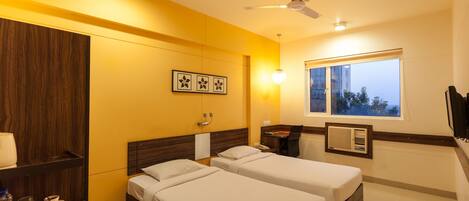 A hotel room with two beds, a wooden wardrobe, a desk with a chair, a window with a view, and a ceiling fan.