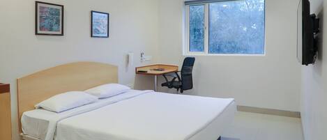 Quarto de hotel com cama, escrivaninha, duas imagens emolduradas, ar condicionado e janela com vista para árvores.