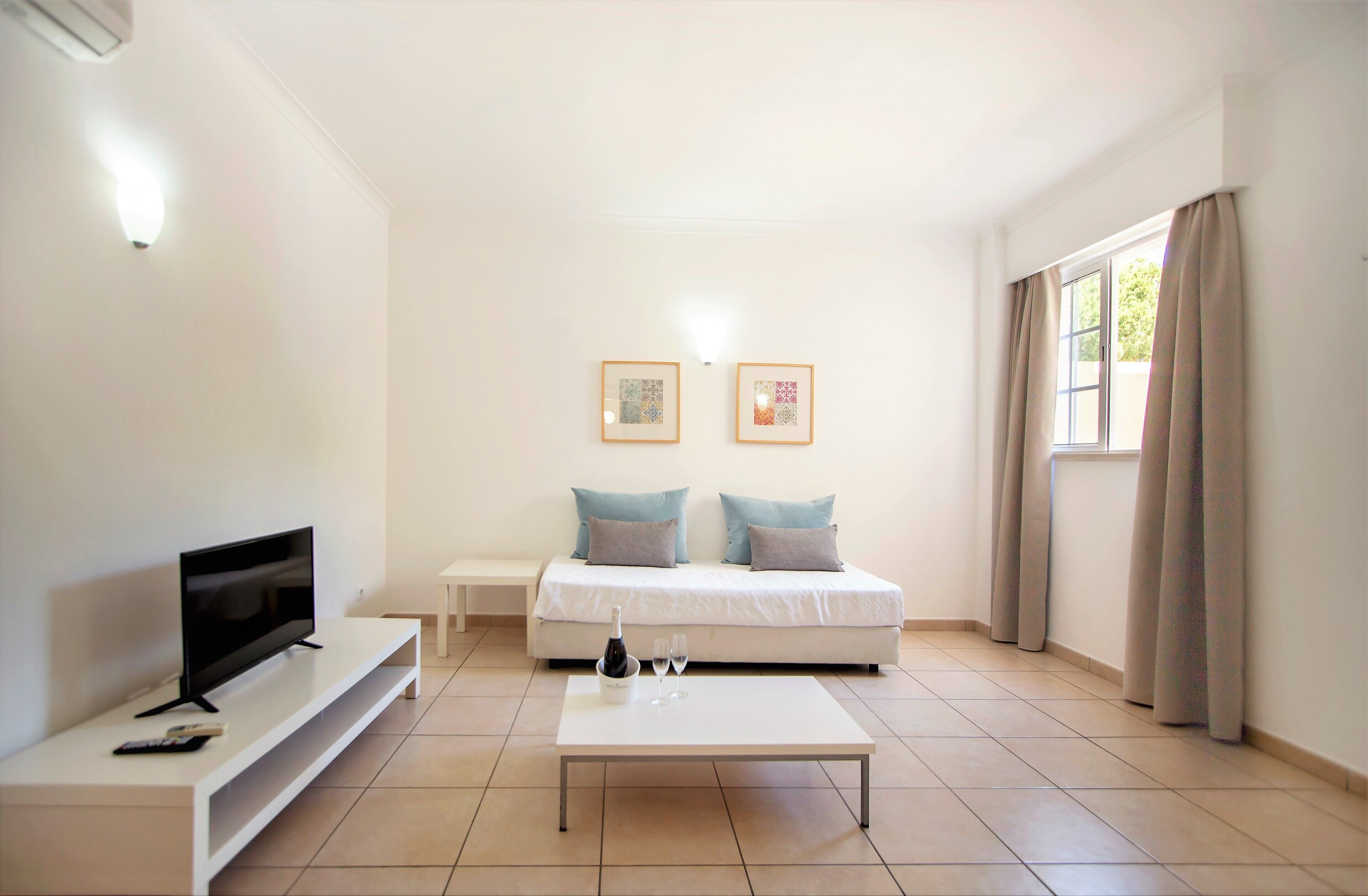 Ein modernes, helles Zimmer mit einem Flachbildfernseher, einem weißen Sofa mit zwei Kissen, einem kleinen weißen Tisch und einem Couchtisch mit Flasche und Gläsern.