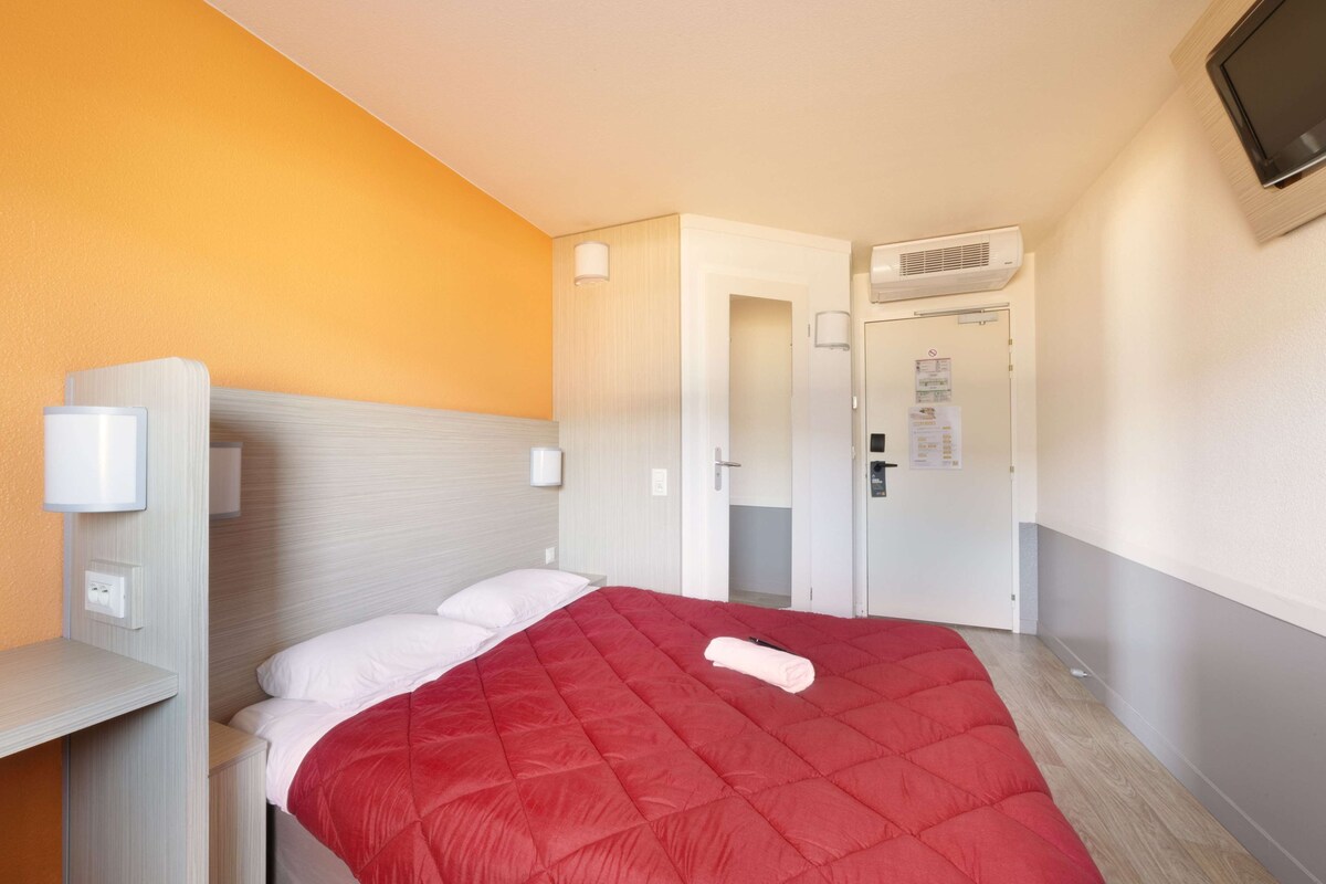 A hotel room with a bed, a bedside table, a wall-mounted TV, and an air conditioning unit.