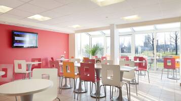 Uma cafeteria bem iluminada, com cadeiras brancas e vermelhas, uma janela ampla e uma televisão de tela plana na parede.