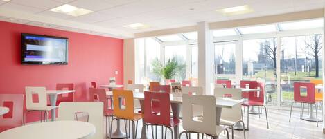 A brightly lit cafeteria with white and red chairs, a large window, and a flat-screen TV on the wall.