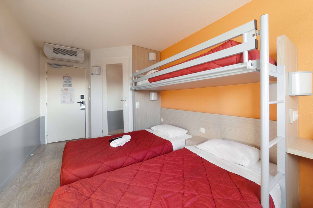 Habitación de hotel con literas, una colcha roja, un almohadón blanco y una puerta con un aviso.