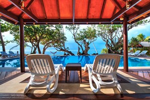Un espace piscine abrité, avec deux chaises longues blanches et une petite table, offrant une vue dégagée sur une piscine aux eaux bleues et sur l’océan.