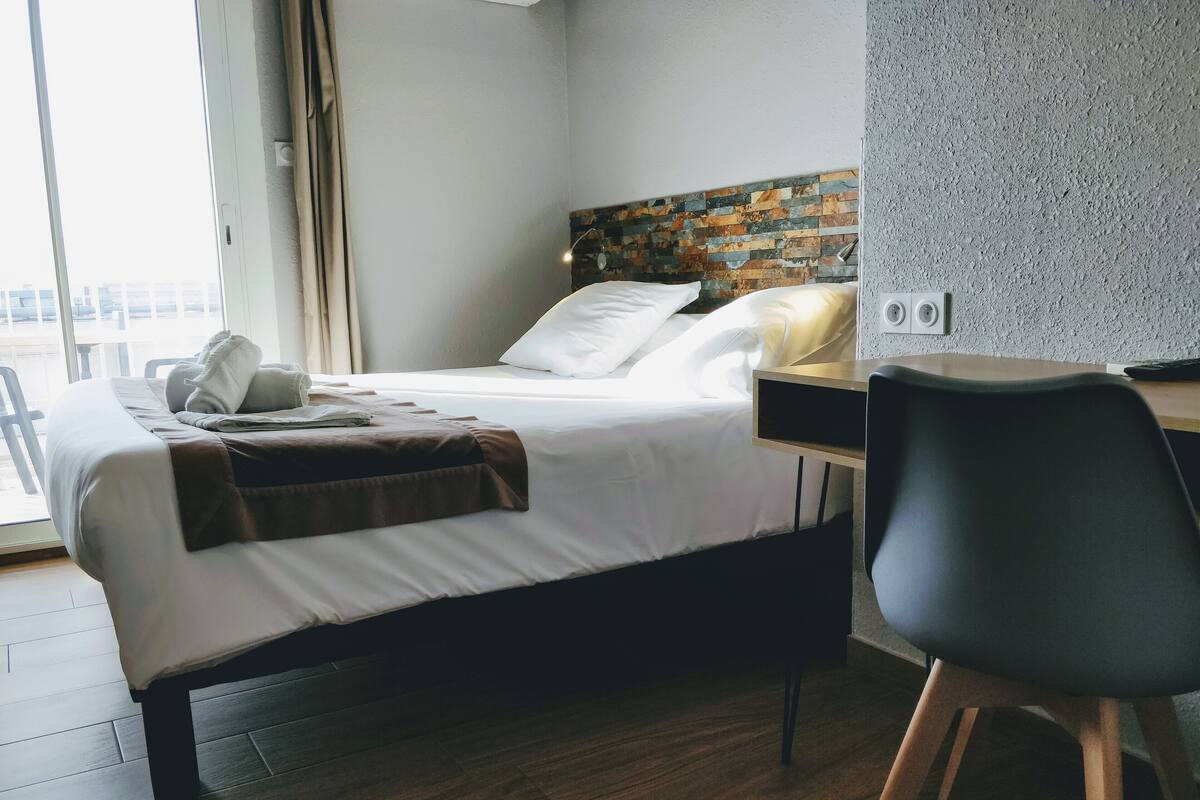 A hotel room with a bed, a desk, a chair, and a view of the city through the window.