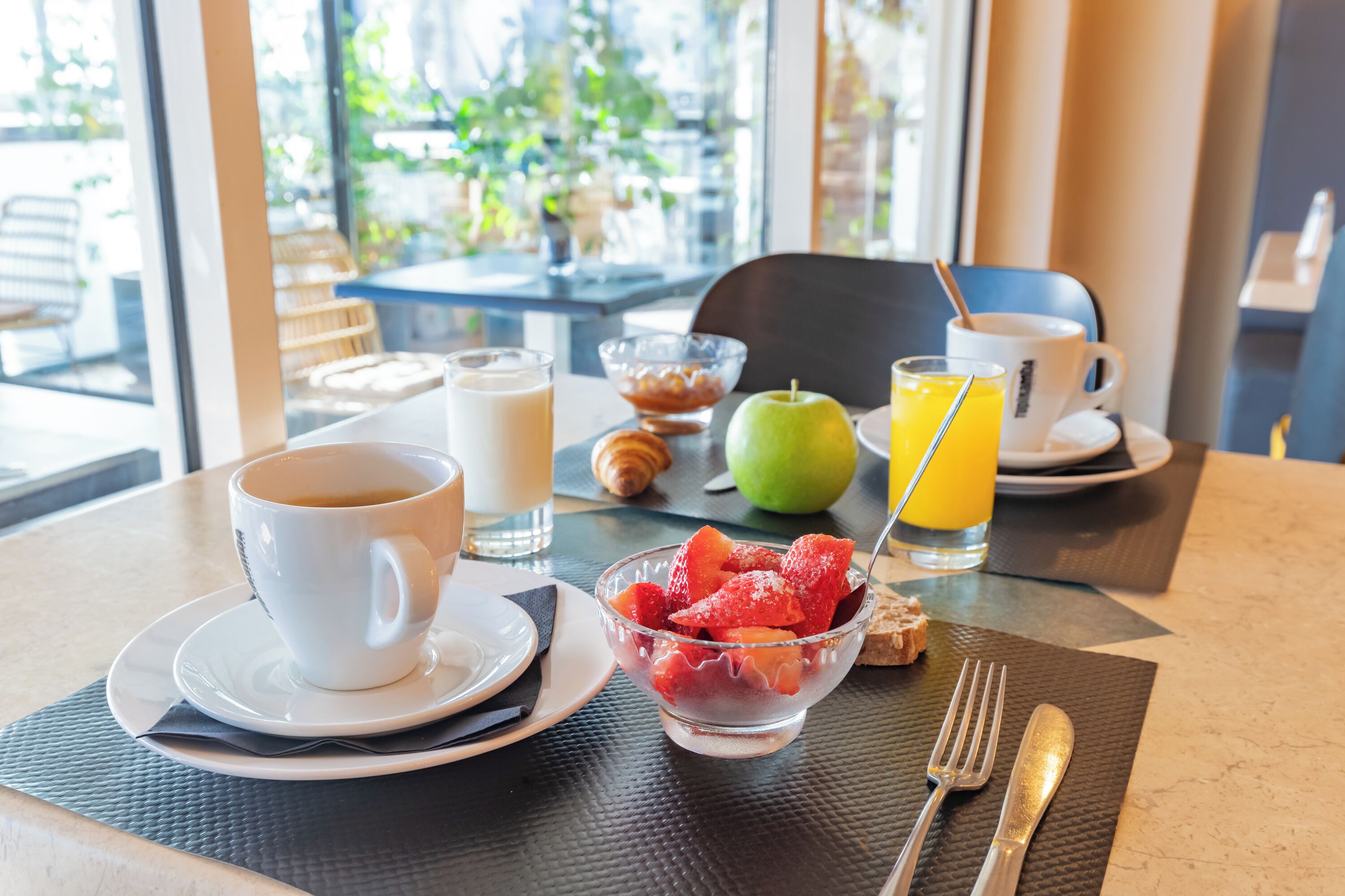 Una mesa de desayuno con una taza de café, un bol de fruta, un vaso de zumo de naranja y croissants.