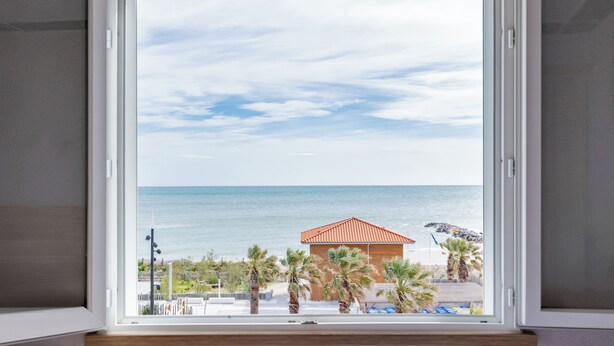 A window with a view of the sea, a building with a red roof, and palm trees. - Mar I Sol (Saint-Cyprien)