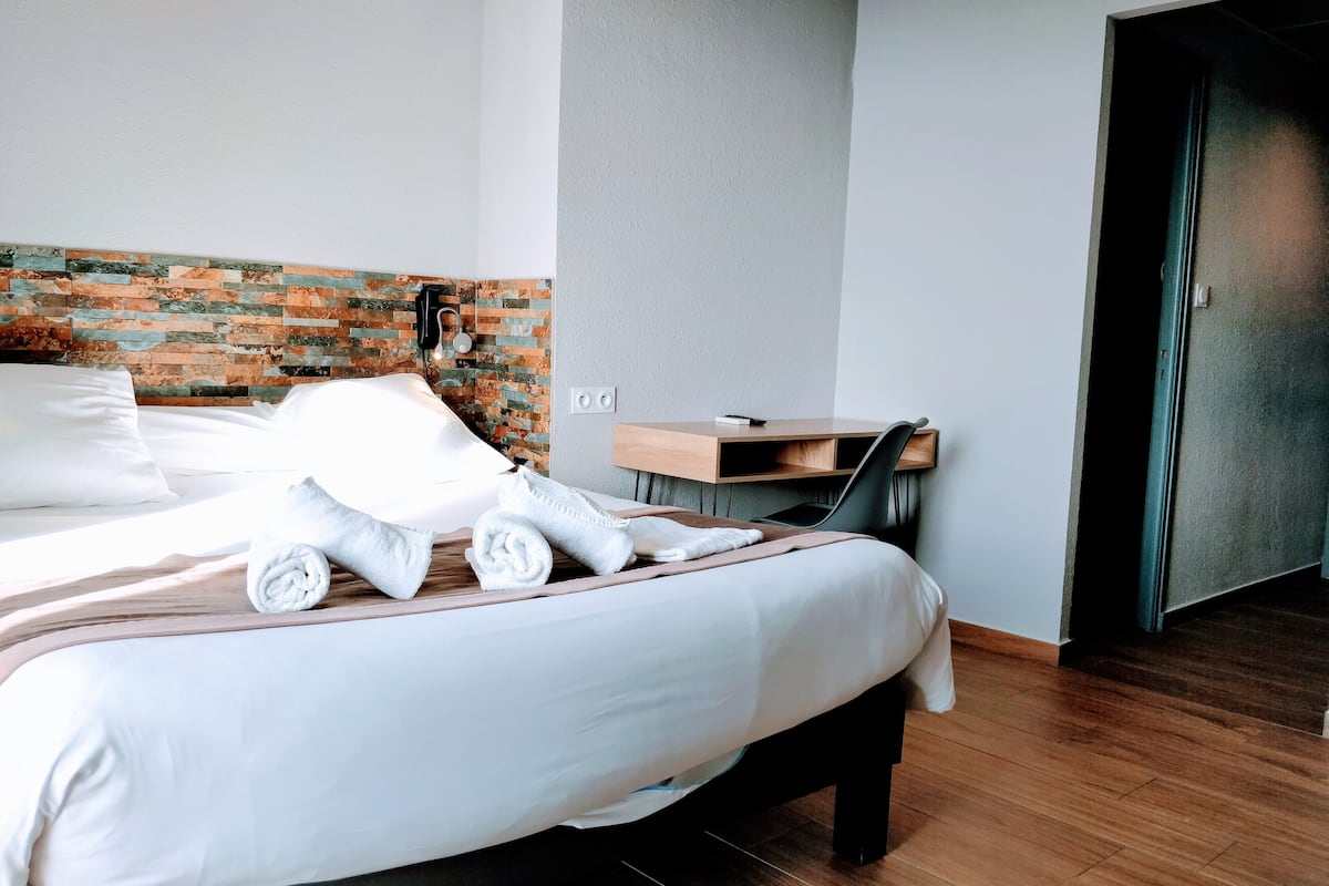 A neatly made bed with white linens, a wooden desk with a chair, and a brick-patterned wall.