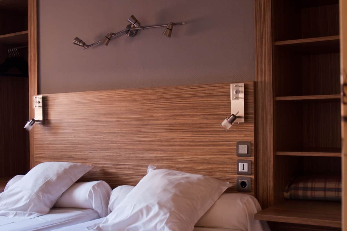 A neatly made bed with white linens and pillows, a wooden headboard with built-in lighting, and a small shelf with a plaid item.