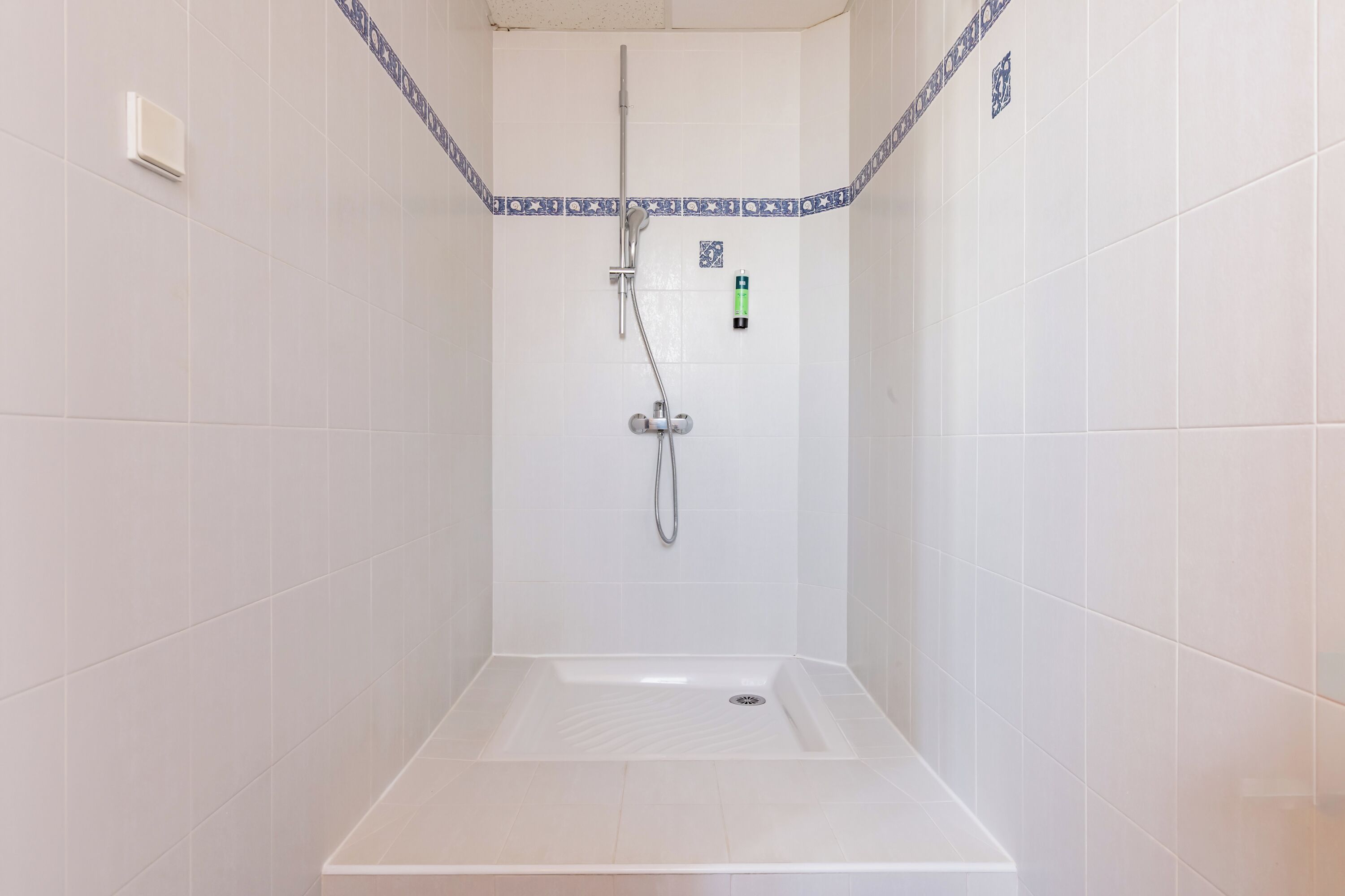 A shower area with a white tiled wall, a showerhead, a handheld shower, and a control panel.