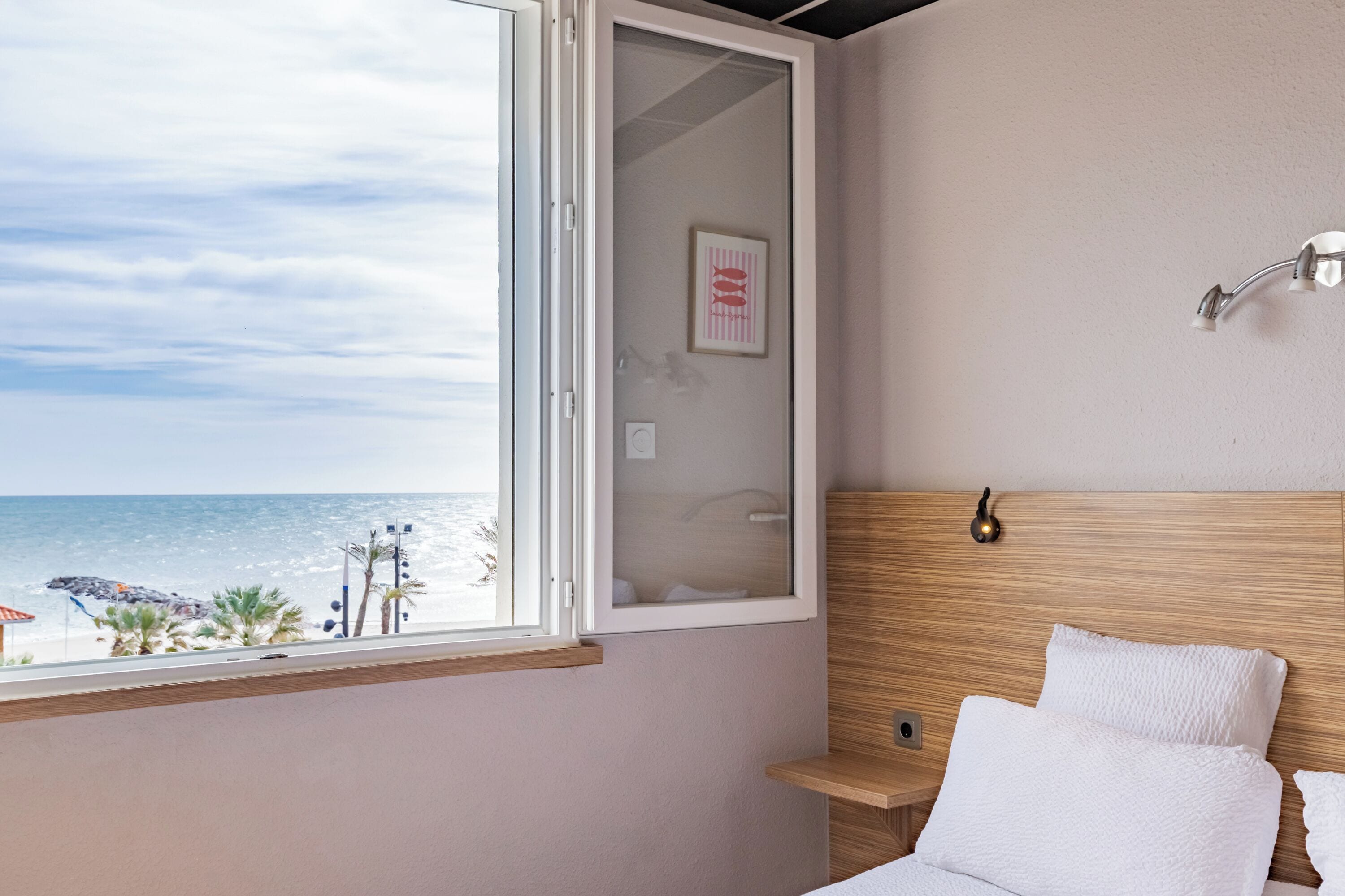 A bedroom with a wooden headboard, a bedside table, a window with a view of the beach, and a framed picture on the wall.