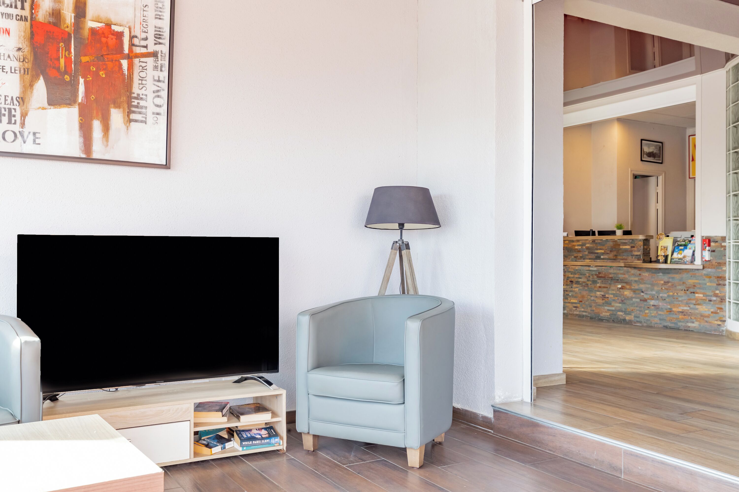 Een woonkamer met een flatscreen-televisie, een lichtblauwe fauteuil, een vloerlamp en een boekenkast.