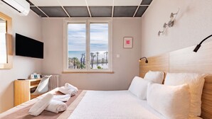 A hotel room with a bed, a TV, a window with a view of the beach, and a desk with a chair. - Mar I Sol (Saint-Cyprien)