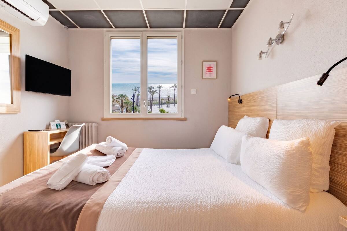A hotel room with a bed, a TV, a window with a view of the beach, and a desk with a chair.
