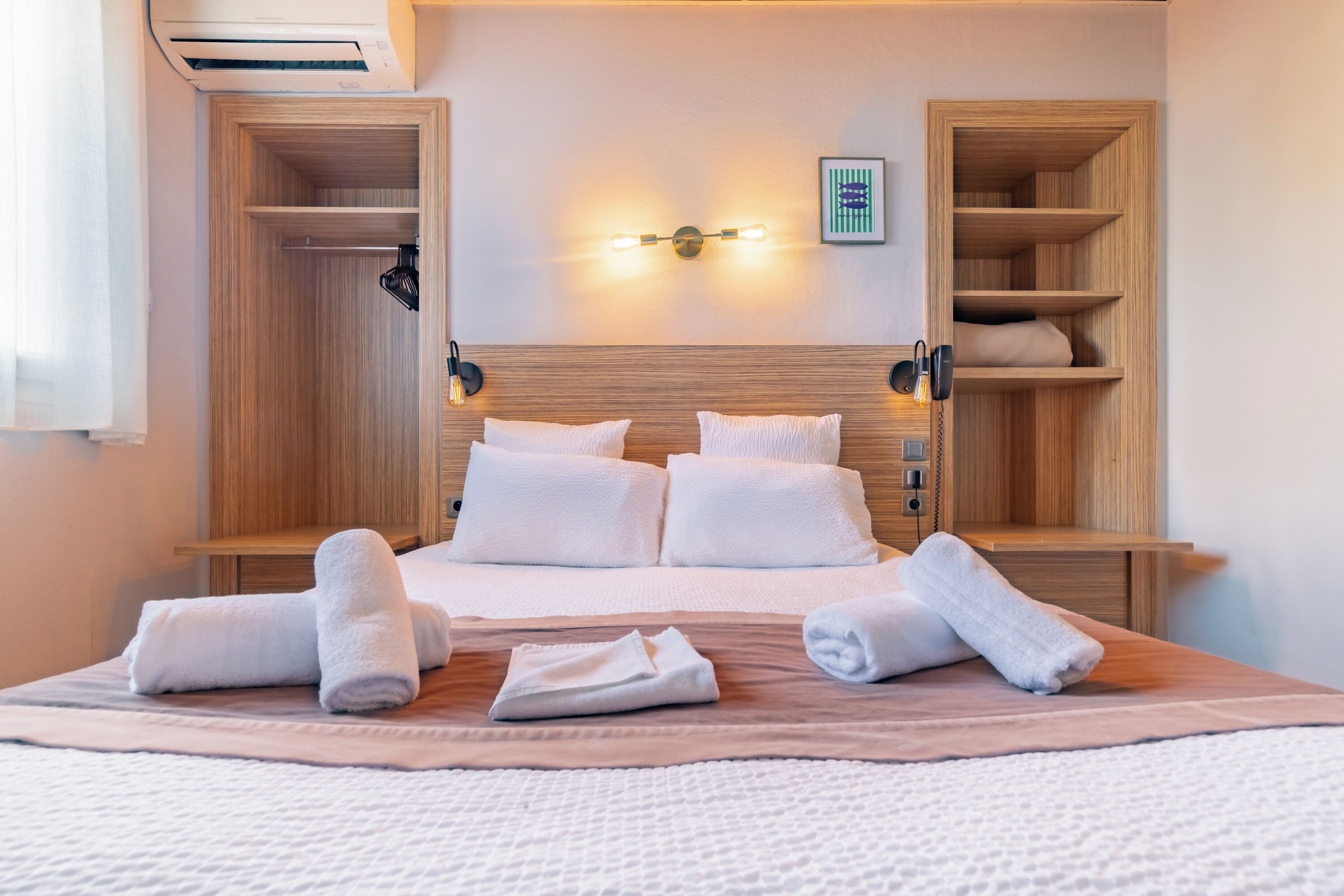 Quarto de hotel com cama, mesa de cabeceira, luminária na parede e estantes de madeira embutidas.