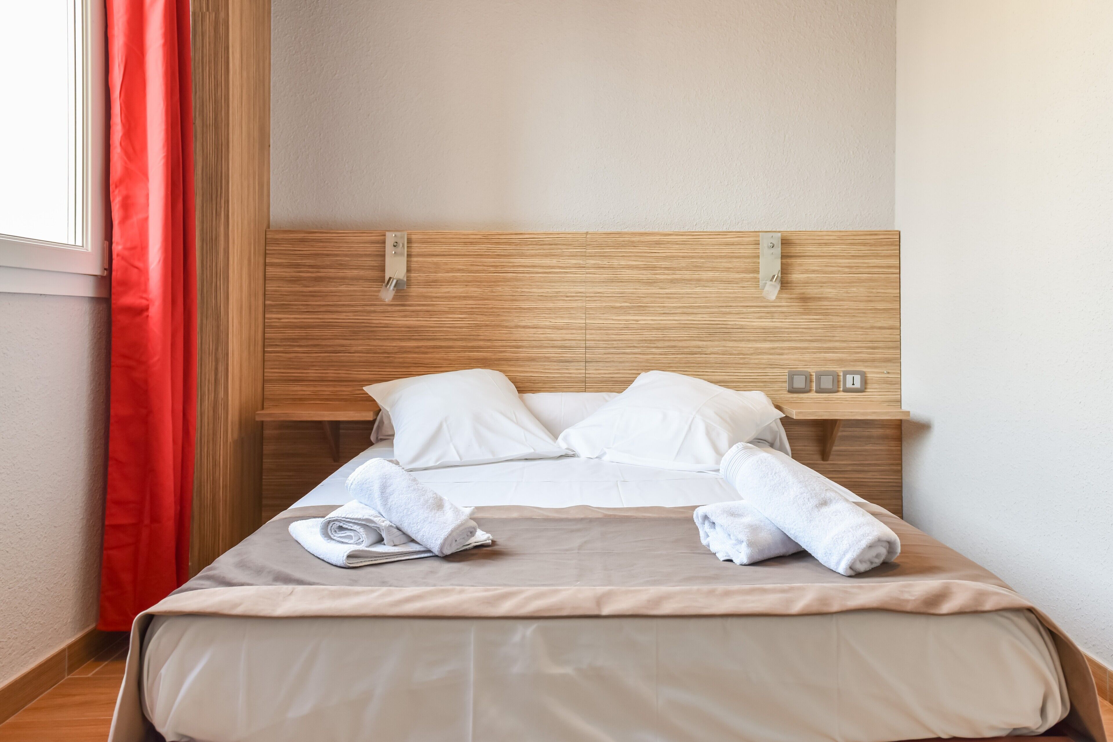 Una cama impecable con sábanas blancas y dos toallas enrolladas, un cabecero de madera y una ventana con cortinas rojas.