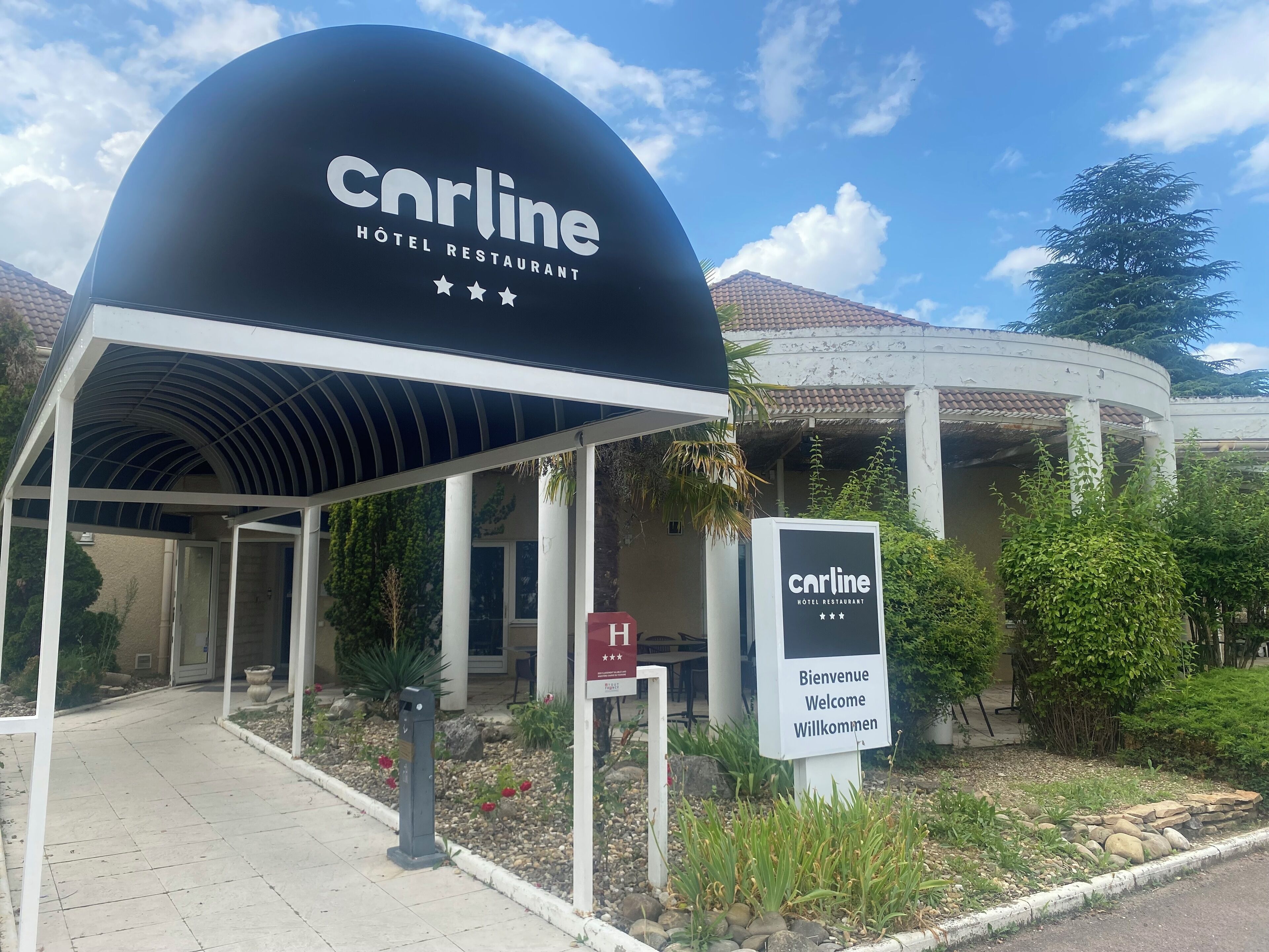 La entrada del restaurante Hotel Carlline, con un corredor cubierto y un cartel que muestra el nombre y un mensaje de bienvenida.
