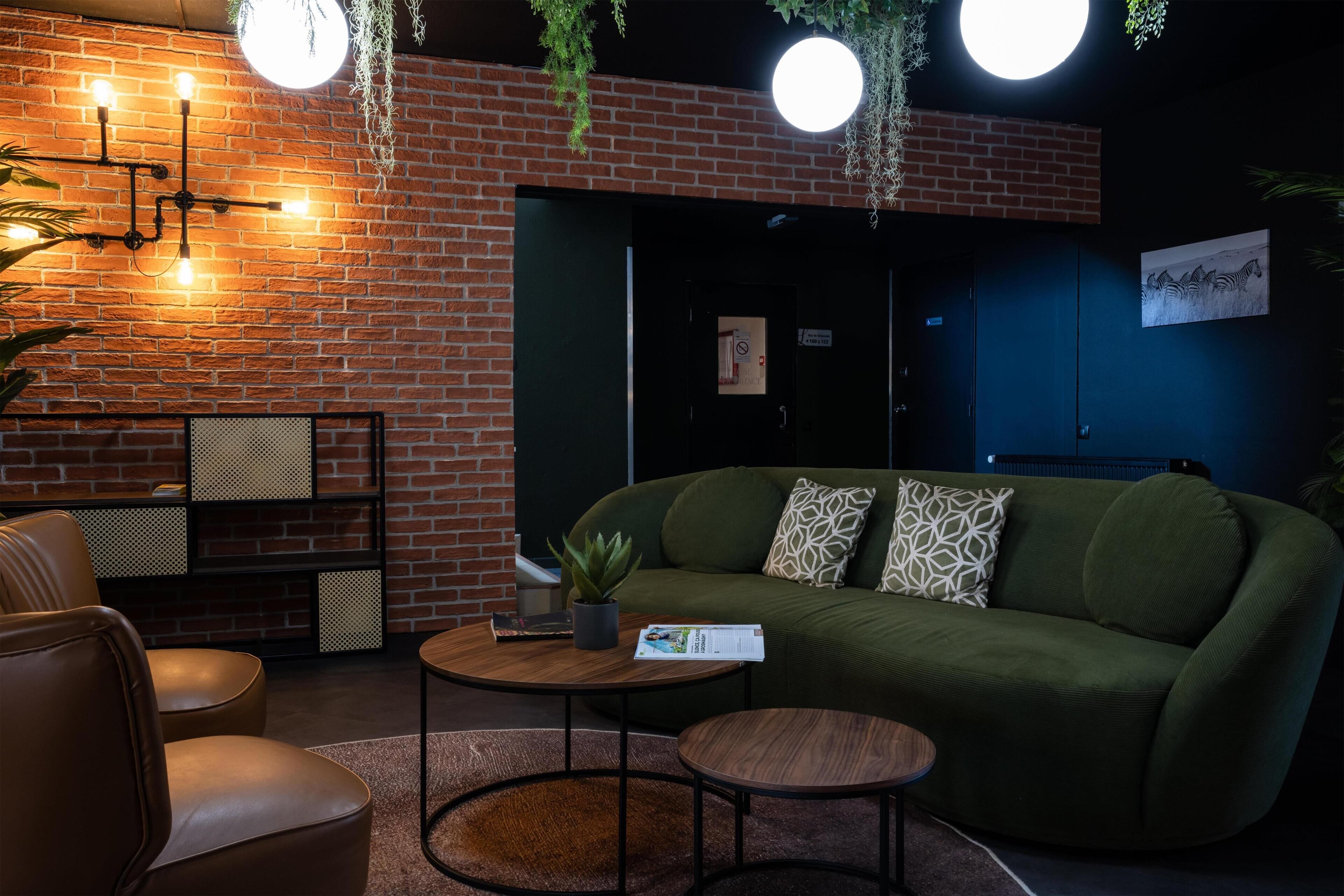 A cozy lounge area with a green sofa, a round wooden table, and a brick wall with hanging lights.