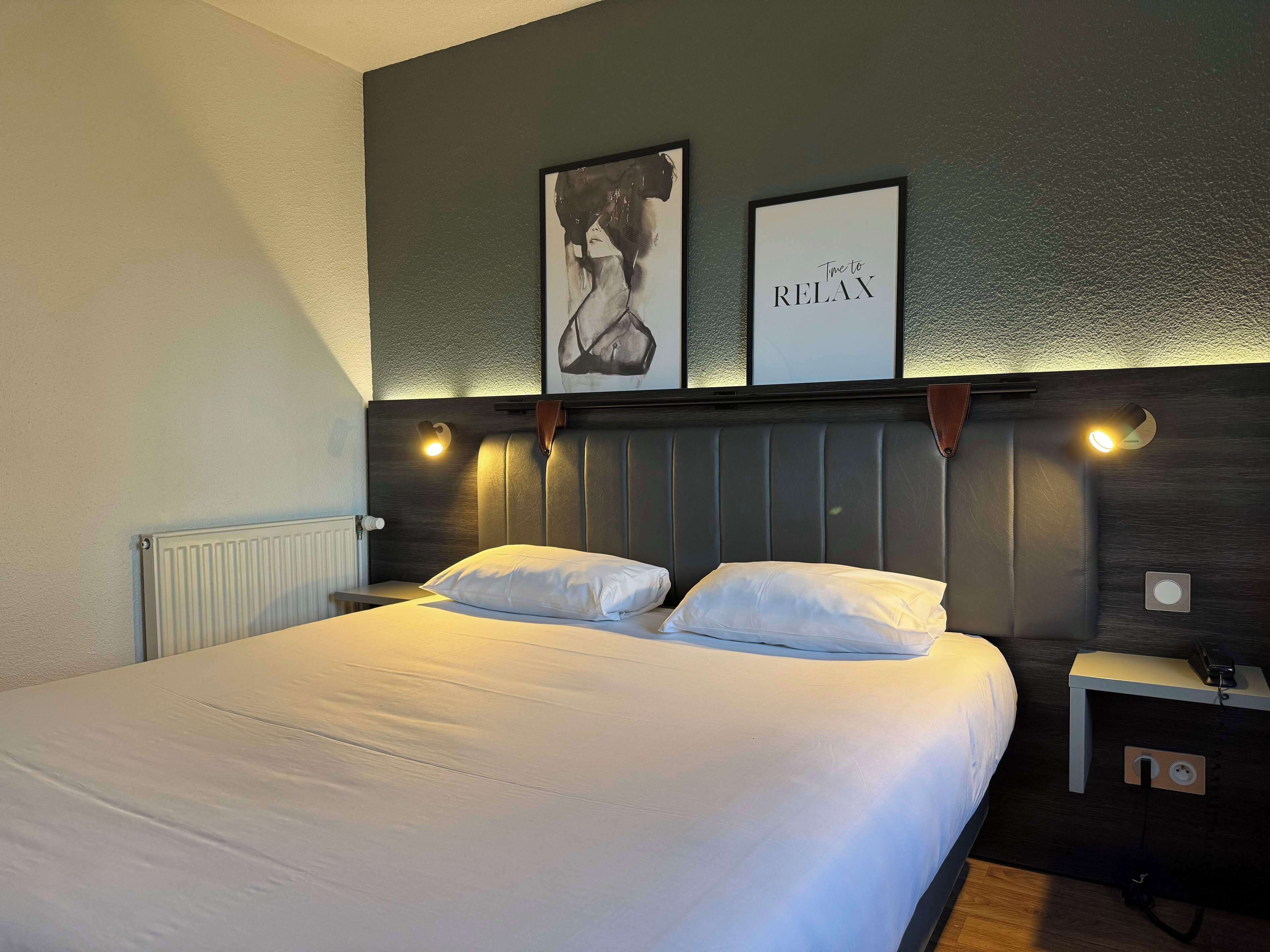 Una cama bien tendida en una habitación de hotel, con cabecera, dos lámparas fijadas a la pared y una obra de arte enmarcada sobre la cama.
