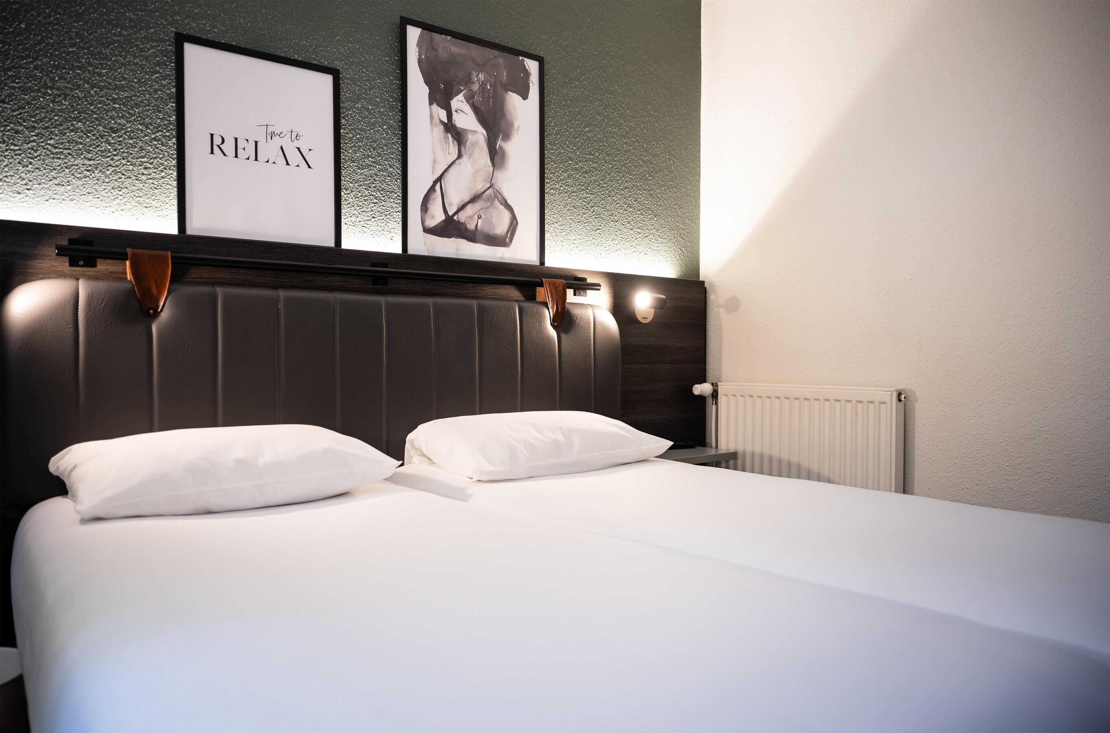 A neatly made bed with white linens and pillows, a dark headboard, and two framed pictures on the wall above.