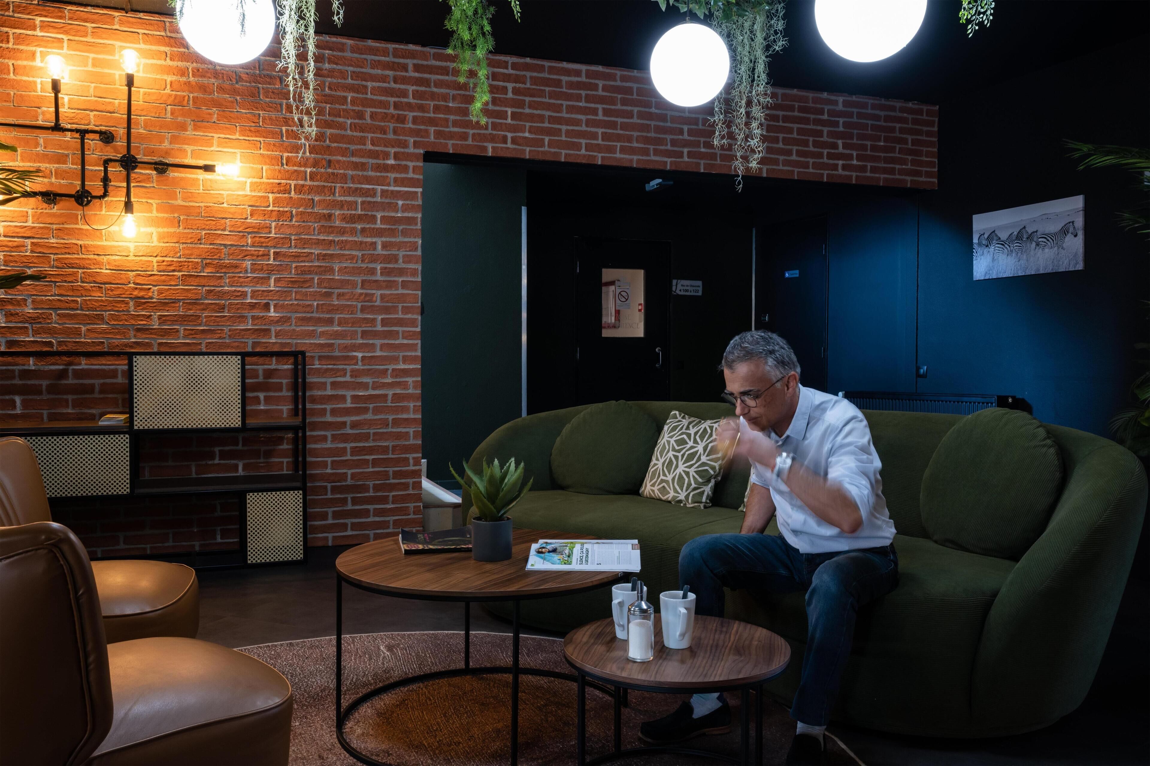 Un área de descanso moderna con pared de ladrillo, luces colgantes, sofá verde y una mesa redonda con una revista y vasos.