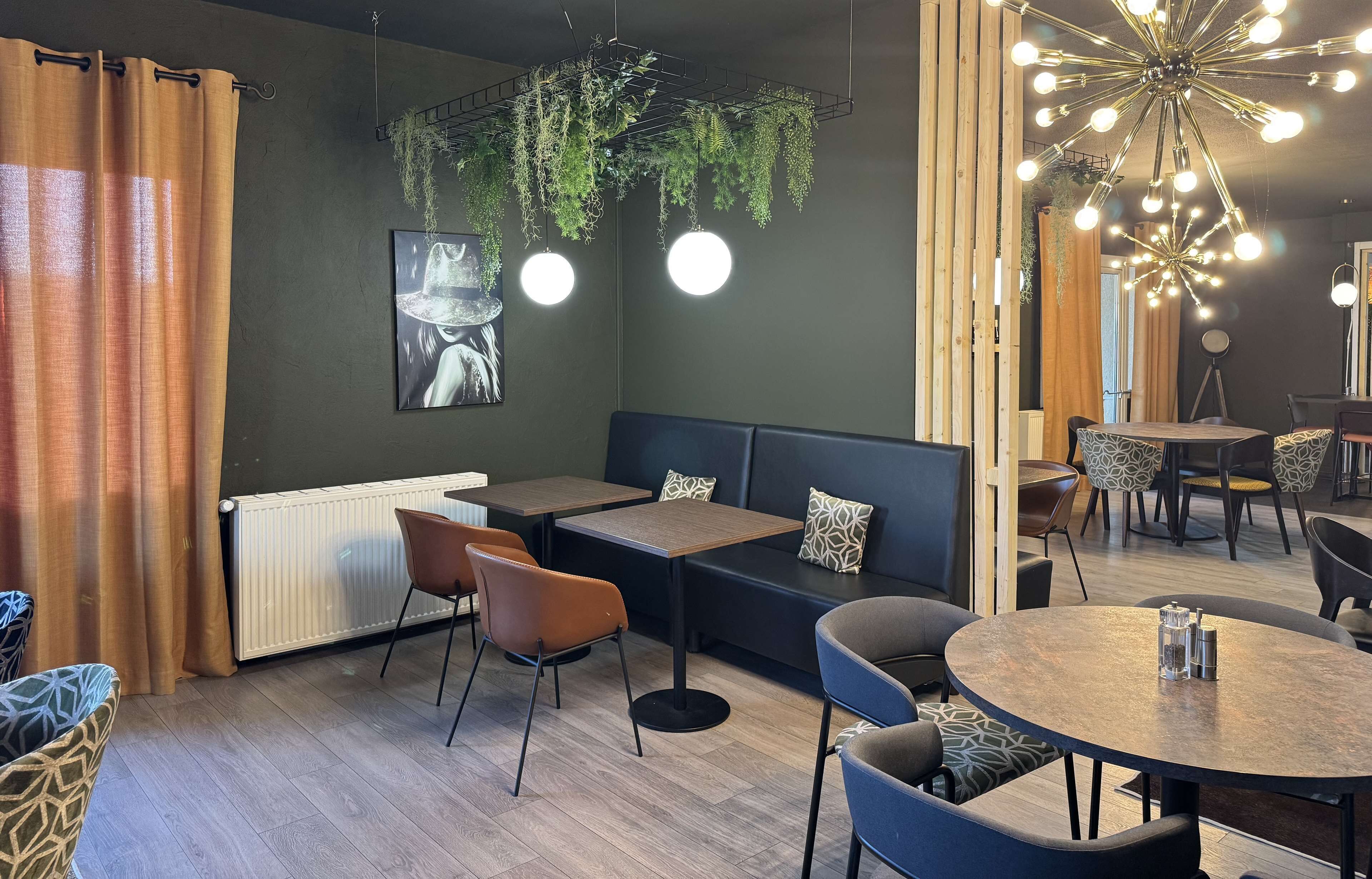 Una cafetería moderna con suelo de madera, asientos negros y marrones, y una pared verde con plantas colgantes.
