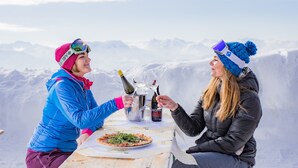 Two people sitting at a table with a pizza and bottles of wine, overlooking a snowy mountain landscape. - Alpenhotel Ensmann (Goestling an der Ybbs)