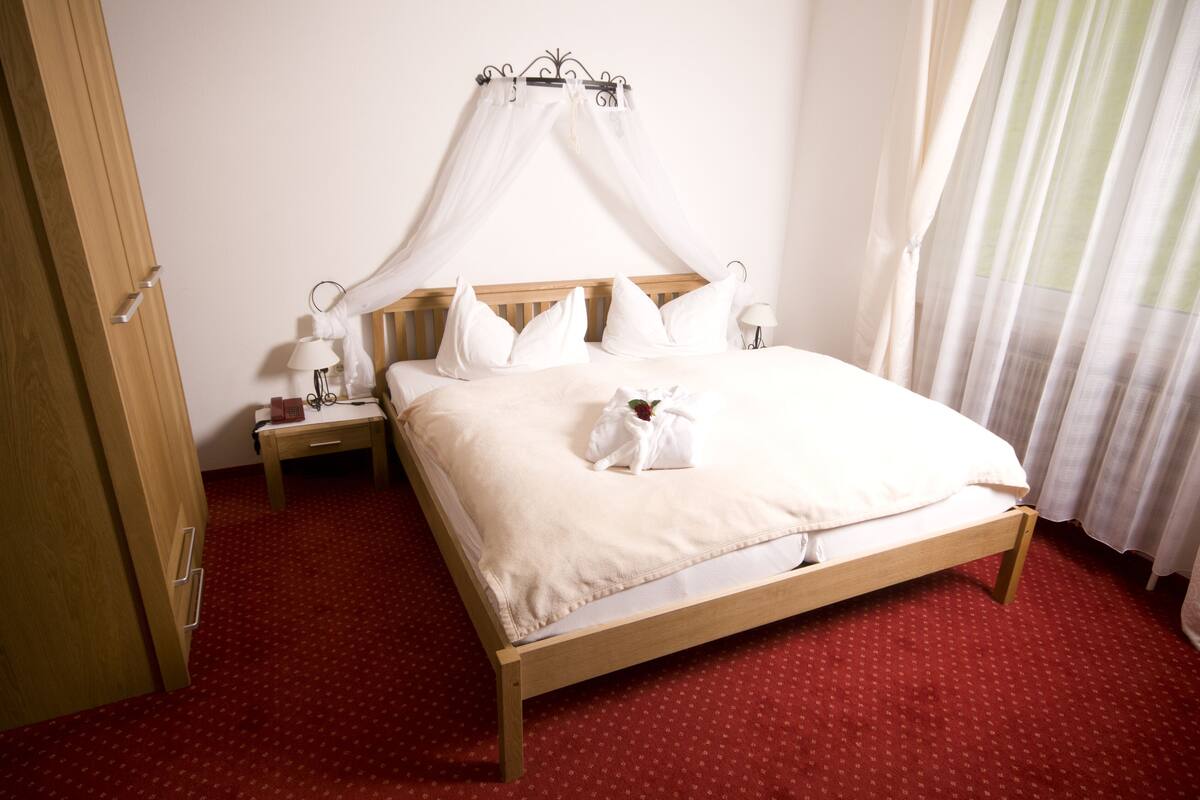 A neatly made bed with a canopy, a bedside table with a lamp, and a red carpeted floor.
