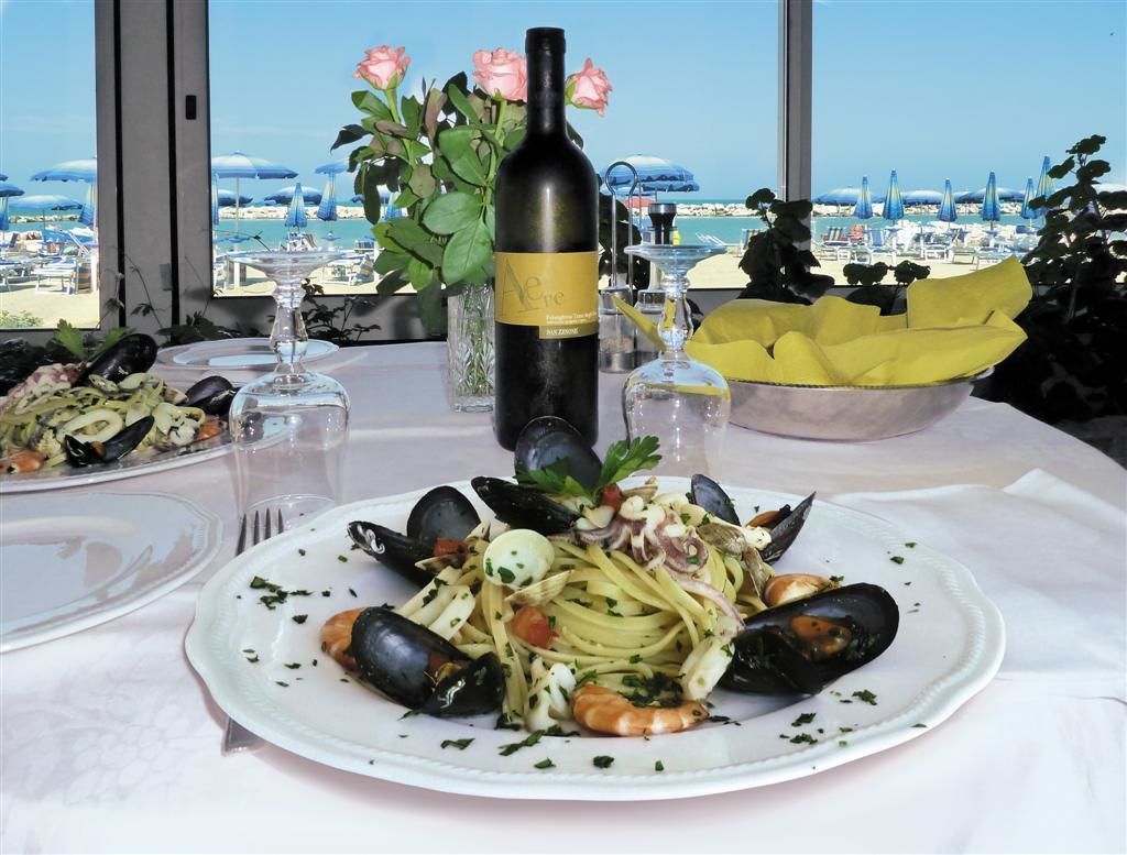Prato de massa com frutos do mar, acompanhado de uma garrafa de vinho e um vaso de flores, sobre uma mesa com vista para a praia.