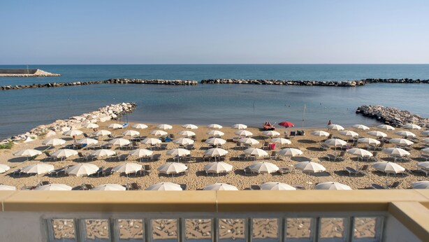A beach with numerous white umbrellas and sun loungers, a rocky breakwater, and calm sea. - Hotel Strand (Montenero di Bisaccia)