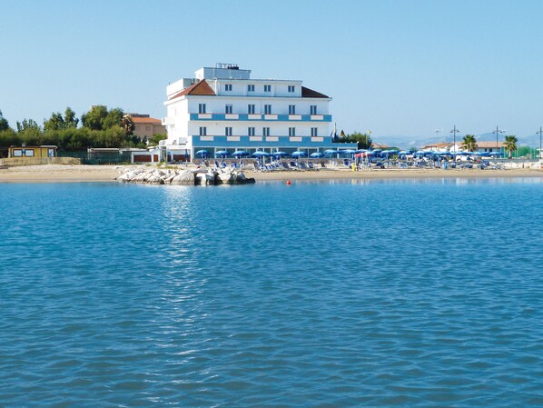 A building with multiple floors, a beach with umbrellas, and a calm body of water.