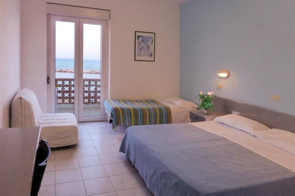 A hotel room with a bed, a chair, a desk, and a balcony with a view of the sea.