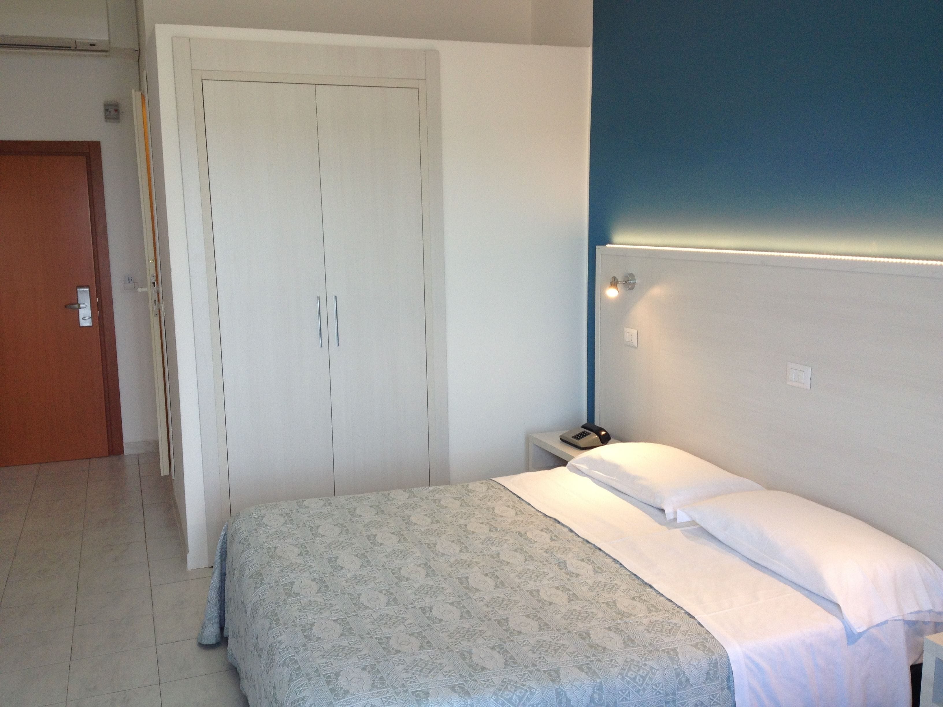 Une chambre à coucher avec un lit, une table de chevet, une porte fermée et un mur muni d’un interrupteur.