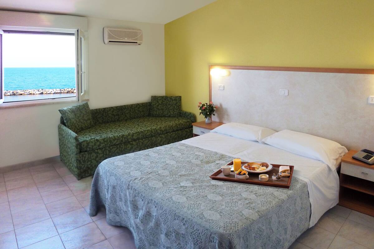 A hotel room with a bed, a green sofa, a bedside table with a phone, and a window with a view of the sea.