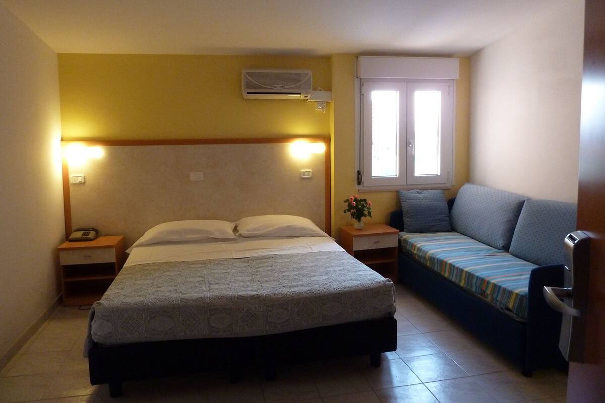 A hotel room with a bed, bedside tables, a sofa, an air conditioning unit, and a window.