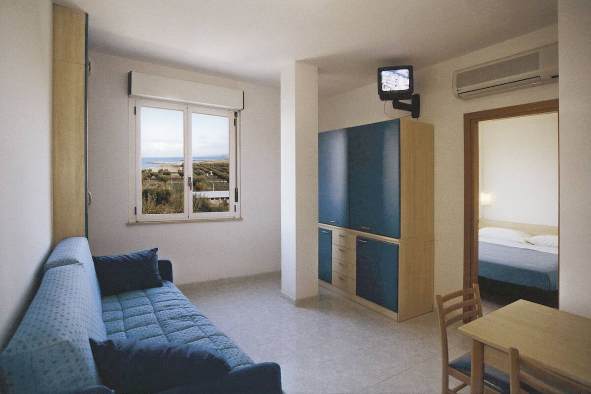 A small room with a bed, a desk, a chair, a TV, and a window with a view of the countryside.